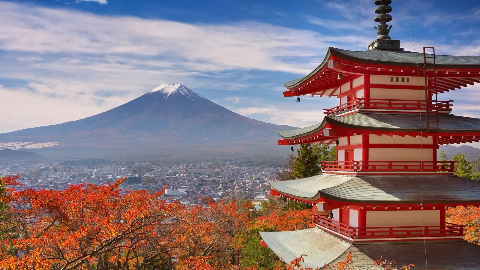 A red five-tiered pagoda on a hillside, with a snow-capped volcano rising behind it and autumn leaves in the foreground.